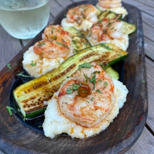 this shows a plate of white grits with cooked shrimp on top, grilled zucchini and creole butter drizzled over the top