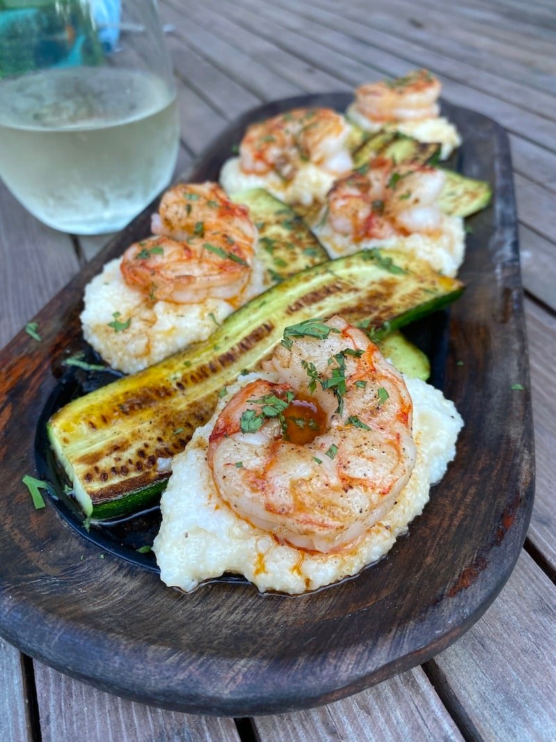 this shows a plate of white grits with cooked shrimp on top, grilled zucchini and creole butter drizzled over the top