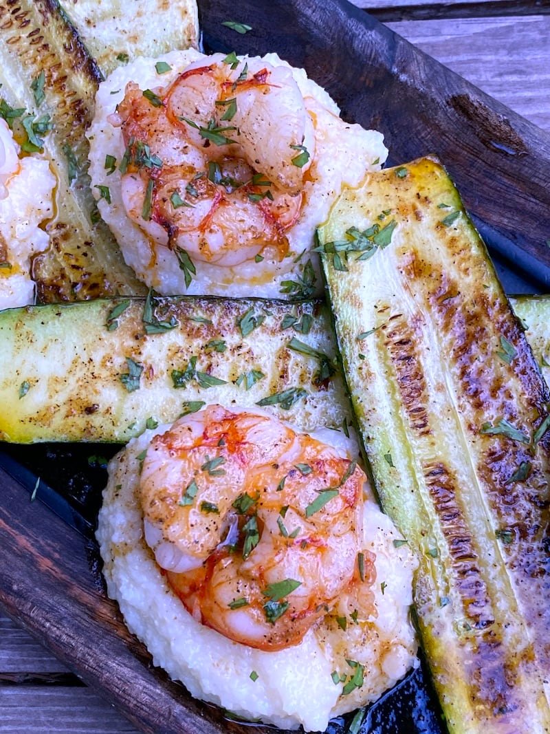 a brown plate that has grits and shrimp with pan fried zucchini drizzled with creole butter