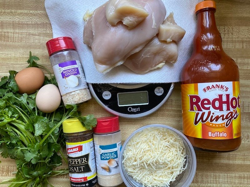 On a counter is all of the ingredients used to make the recipe. Chicken breast, cilantro, eggs, seasonings, hot sauce and Paremsan cheese