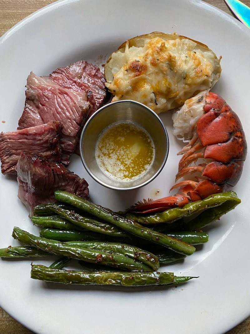 this is a picture of a white plate that has sliced beef, lobster tail, green beans and twice baked potatoes